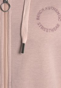 Sudadera con cremallera en color rosa, fabricada en un tejido texturizado, que cuenta con una cremallera plateada, cordones y un logotipo en relieve que dice "BENCH AUTHENTIC STREETWEAR."