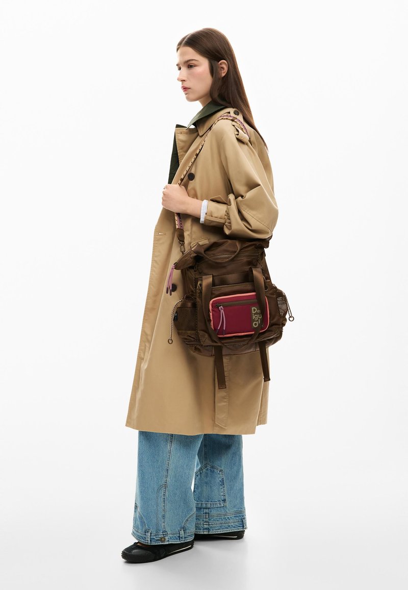 Femeie purtând o jachetă trench bej și blugi cu picioare evazate poartă un rucsac mare maro cu buzunar roz pe partea din față pe un umăr.