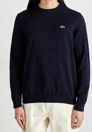 Person iført en mørkeblå langærmet crewneck-trøje med en lille grøn krokodillelogo på venstre bryst og off-white bukser.