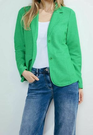 Femme portant un blazer vert, un haut blanc et un jean bleu, une main dans la poche, debout devant un fond clair uni.