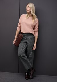 Maglione rosa, pantaloni a gamba larga grigio a scacchi, borsa bordeaux e stivaletti coordinati. Modella in posa su uno sfondo scuro.