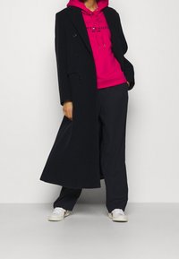 Długi czarny płaszcz dwurzędowy na jasnoróżowej bluzie z kapturem i sznurkami, w połączeniu z ciemnymi spodniami i białymi trampkami.