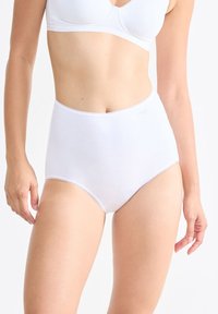 Vrouw in een witte high-waisted slip en witte bh, staande tegen een effen lichte achtergrond met ontspannen armen aan haar zij.