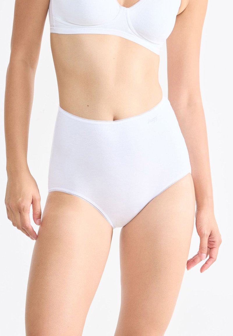 Vrouw in een witte high-waisted slip en witte bh, staande tegen een effen lichte achtergrond met ontspannen armen aan haar zij.
