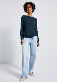 Türkise Langarmbluse kombiniert mit hellblauen, weiten Jeans. Strickmaterial, enganliegende Bündchen und lockerer Ausschnitt. Schlichte beige Schuhe.