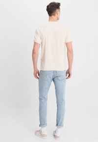 En man står vänd bort med en kortärmad beige skjorta, ljusblå jeans med uppvikta ben, vita strumpor och rosa Adidas-sneakers.