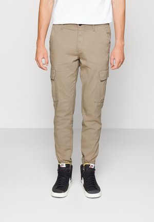 Man draagt beige cargobroek met zijzakken en zwarte Nike hoge sneakers, staand tegen een effen lichte achtergrond.