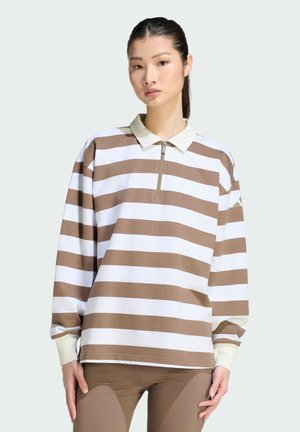 Jonge vrouw die een bruin-wit breed gestreept poloshirt met lange mouwen en bruine legging draagt, staand tegen een effen achtergrond.