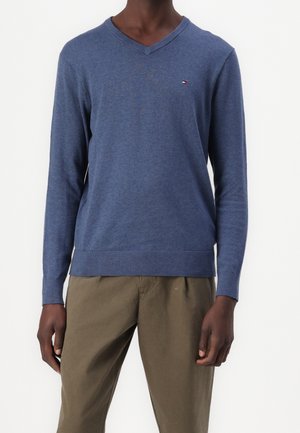 Homme portant un pull bleu à col en V avec un petit logo sur la poitrine et un pantalon vert olive, debout devant un fond blanc.