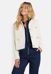 Chaqueta de tweed blanca corta con botones dorados y dos bolsillos frontales. Lleva puesta sobre un mono azul marino con cremallera y detalle de cinturón.