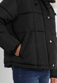 Svart quiltad vinterjacka med silverfärgade tryckknappar, sidoficka, ribbad mudd och pälskant på huvan, bärs med blå jeans.