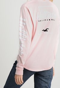 Maglietta a maniche lunghe rosa con logo "Hollister" e grafica di un gabbiano, con testo bianco sulle maniche e vestibilità comoda. Tessuto di cotone liscio.