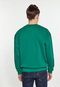 Groene sweatshirt met lange mouwen, geribbelde halslijn en manchetten, met een matte afwerking. G gedragen met donkerblauwe jeans en zichtbare stikseldetails.