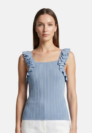 Giovane donna che indossa un top smanicato a coste azzurro chiaro con spalline arricciate e pantaloni bianchi, in piedi davanti a uno sfondo semplice.