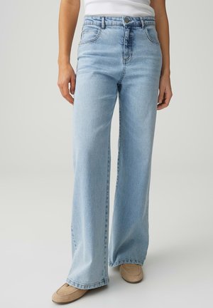 Person trägt hellblaue, weit geschnittene Jeans mit hohem Bund, kombiniert mit beigen Wildleder-Loafers und einem weißen ärmellosen Oberteil.