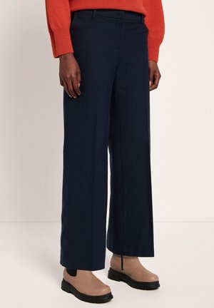 Person wearing navy wide-leg pants, beige ankle boots with black soles, and an orange sweater standing on a light floor and wall background.