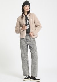 Fleece bomberjack in lichtbeige met een rits aan de voorkant, gecombineerd met hoog opgesneden vervaagde grijze jeans en zwarte sneakers met witte zolen.