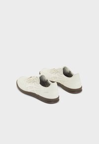 Světlé béžové tenisky s texturovaným koženým svrškem, obsahující bílé pruhy, kulaté šněrovadla a hnědou gumovou podrážku. Doplňky zahrnují značení na patě.