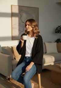 Czarny dziergany kardigan na białej koszulce, siedzący na drewnianym stołku, trzymający białą filiżankę, w tle niebieskie dżinsy i beżowa sofa.