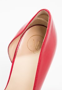 Chaussure à talon haut en cuir rouge avec intérieur beige et lettres du logo dorées M et S à l’intérieur de la zone du talon.