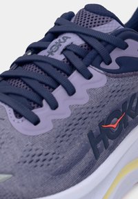 Μωβ και γκρι παπούτσι τρεξίματος με πάνω μέρος από δίχτυ, χρωματικούς κορδόνια, ενισχυμένες θηλιές, λογότυπο HOKA και μαλακή λευκή σόλα με κίτρινη λεπτομέρεια.