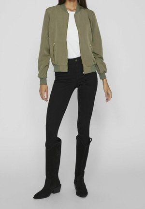 Kvinde iført en olivengrøn bomberjakke med lynlåslommer, hvid skjorte, sorte højtaljede bukser og sorte knælange støvler mod en grå baggrund.