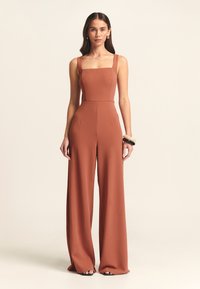 Terracotta kleurige jumpsuit met een vierkante halslijn, brede benen en verstelbare bandjes. De textuur van de stof en subtiele zijzakken verbeteren het ontwerp.