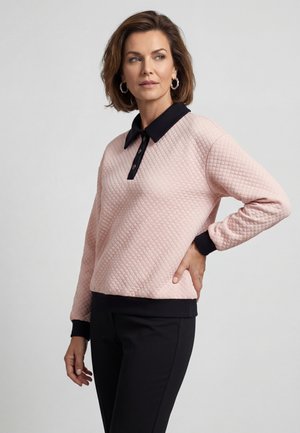 Donna con top trapuntato rosa a manica lunga con collo e polsini neri, pantaloni neri, mano sul fianco, guarda di lato contro uno sfondo uniforme.