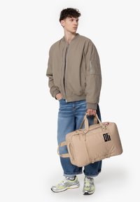 Beige bomberjacka med en ficka med dragkedja, blå jeans och en beige duffelväska med handtag. Sneakers med silver- och neon-design.