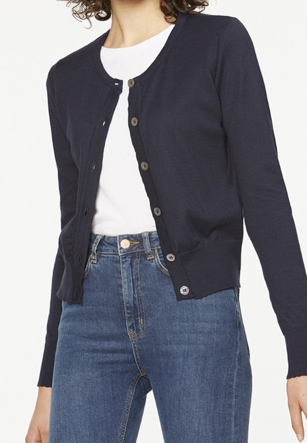 Femme portant un cardigan bleu marine boutonné sur un t-shirt blanc, associé à un jean bleu taille haute sur fond blanc.