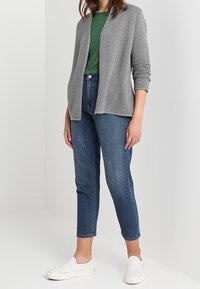 Cardigan ouvert côtelé gris sur un haut vert, associé à un jean bleu denim longueur cheville et des chaussures blanches à enfiler. Design simple et texturé.