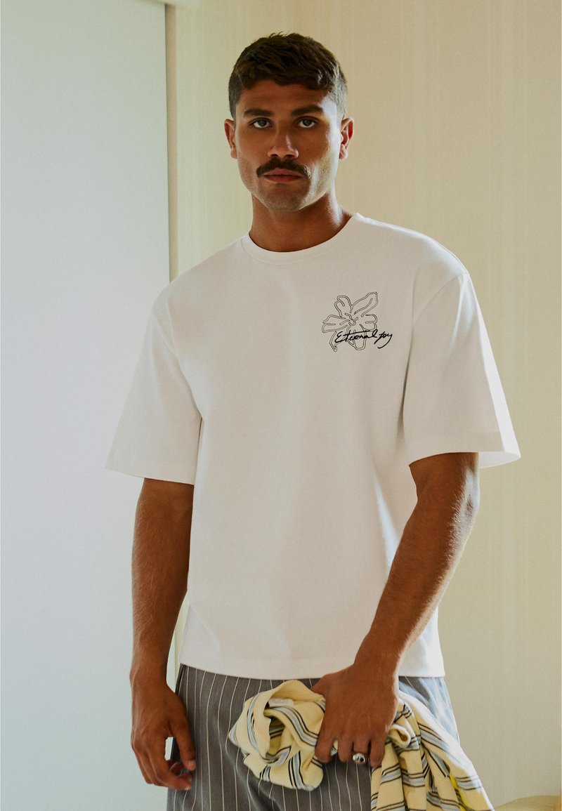 Homme avec moustache portant un t-shirt graphique blanc et un pantalon rayé, tenant un tissu rayé dans un cadre intérieur neutre.