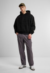 Hoodie en polaire noir avec une poche kangourou, associé à un pantalon en velours côtelé gris et des baskets blanches avec des rayures noires.