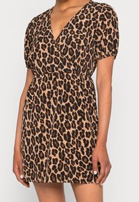 Femme portant une robe à manches courtes, col en V, imprimé léopard avec une taille élastique, visible du cou jusqu'à mi-cuisse sur un fond uni.