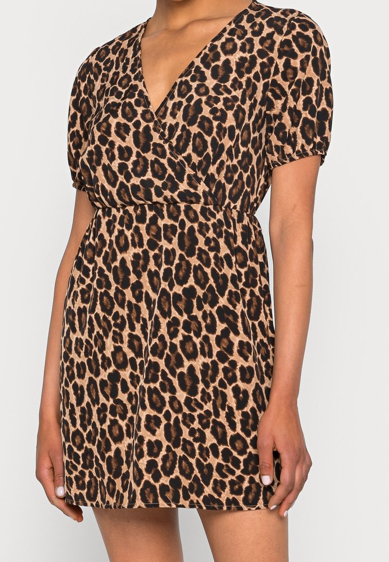 Femme portant une robe à manches courtes, col en V, imprimé léopard avec une taille élastique, visible du cou jusqu'à mi-cuisse sur un fond uni.