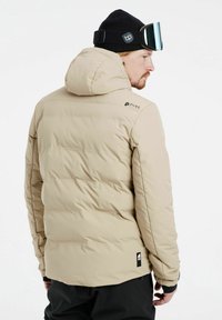 Beige Daunenjacke mit Kapuze, ausgestattet mit einem gesteppten Design, elastischen Bündchen und einem Logo auf der linken Schulter. Strukturiertes Material mit matter Oberflächenbeschaffenheit.