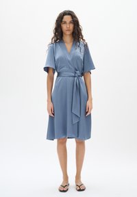 Blaues Midikleid mit Wickeldesign, kurzen Ärmeln, einem gebundenen Taillengürtel und einer glatten Textur. Getragen mit einfachen schwarzen Flip-Flops.