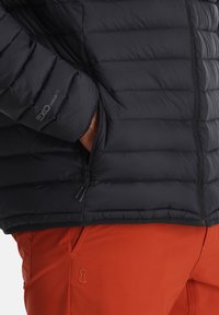 Zwarte pufferjack met horizontale stiksels, met een ritszak aan de zijkant en een logo op de mouw. Gecombineerd met oranje broek.
