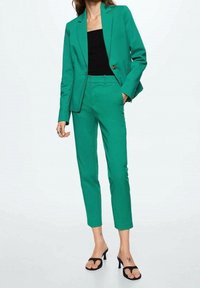 Costume vert sur mesure avec un blazer à un seul bouton, un pantalon slim et un haut noir. Le matériau semble lisse avec une couleur vive et unie.