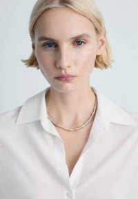 Sølv kæde med tekstureret, perle-designet båret over en hvid skjorte. Kort, lige blond hår og naturlig makeup.