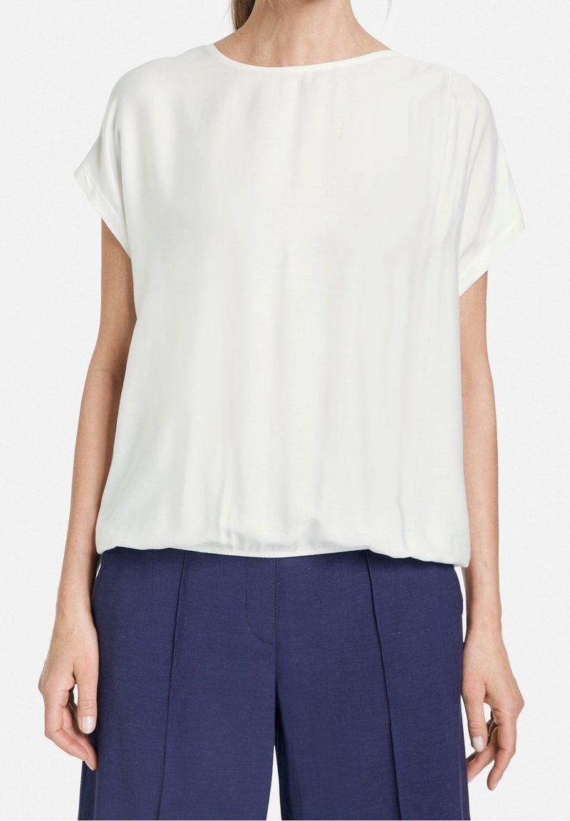 Femme portant un chemisier blanc ample à manches courtes et un pantalon bleu marine taille haute, debout, les bras détendus le long du corps.