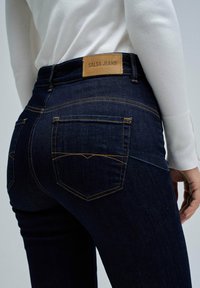 Vaqueros de mezclilla azul oscuro con costuras amarillas y una etiqueta de cuero marrón claro que dice "SALSA JEANS", combinados con una blusa de manga larga blanca.