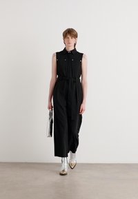 Mono negro sin mangas con cintura ceñida por un cinturón, con detalles plateados en el cuello y los bolsillos. El modelo sostiene un bolso de mano plateado.