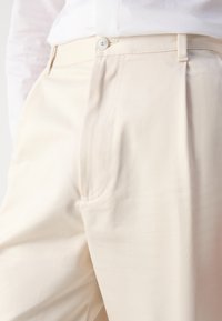 Pantalones de algodón beige con frente plano, bolsillos laterales y cierre de botón. Textura suave y ajuste entallado con detalles de costura sutiles.