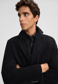 Cappotto di lana nero con collo alto, texture morbida, grandi rever e dettaglio con tre bottoni. Indossato sopra una camicia nera con bottoni.