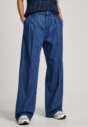 Personne debout portant un pantalon en denim bleu ample avec des plis et des baskets blanches, les mains dans les poches, sur fond uni.