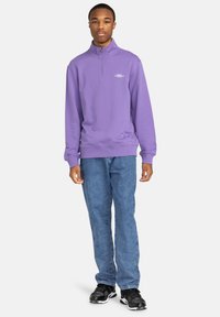 Felpa viola con zip e colletto alto, con un logo sottile. Indossata con jeans di denim blu chiaro e scarpe da ginnastica nere.