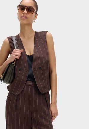 Kvinde iført brun pinstripe ærmeløs blazer og matchende bukser, sort top under, bærer sort håndtaske og solbriller.