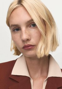 Joven mujer con cabello rubio corto, ojos azules y pequeñas pecas, vestida con una camisa beige con cuello y una chaqueta color óxido.