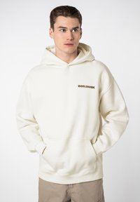 Helle Creme Kapuzenjacke aus weichem Stoff, mit einer Kängurutasche und einem kleinen braunen "WORLDWIDE"-Logo auf der linken Brustseite.